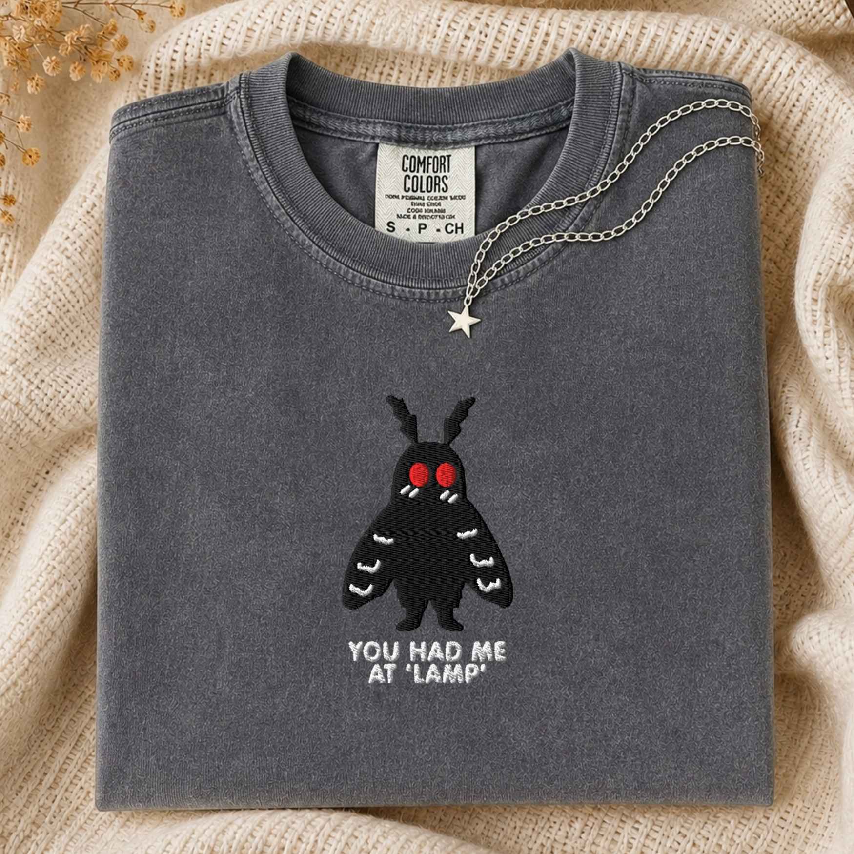 A folded slate gray t-shirt with a black moth graphic and the text 'YOU HAD ME AT "LAMP"' on the front, styled on a beige knit blanket with a silver necklace draped over the collar.