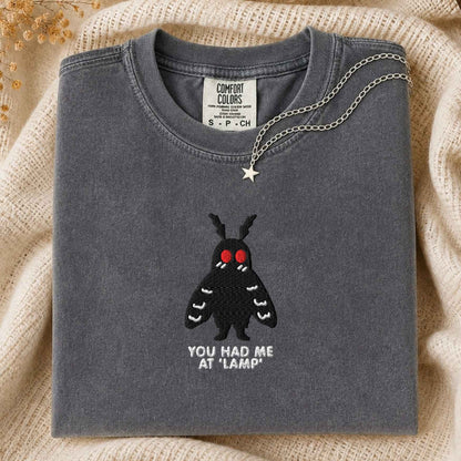 A folded slate gray t-shirt with a black moth graphic and the text 'YOU HAD ME AT "LAMP"' on the front, styled on a beige knit blanket with a silver necklace draped over the collar.