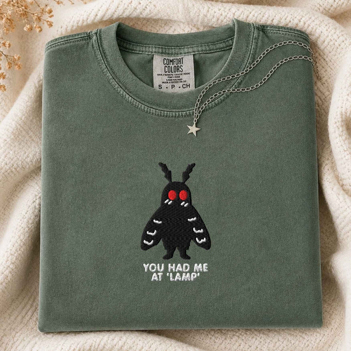 A folded sage green t-shirt with a black moth graphic featuring red eyes and the text 'YOU HAD ME AT "LAMP"' underneath, styled on a cream knit blanket with a silver star necklace.
