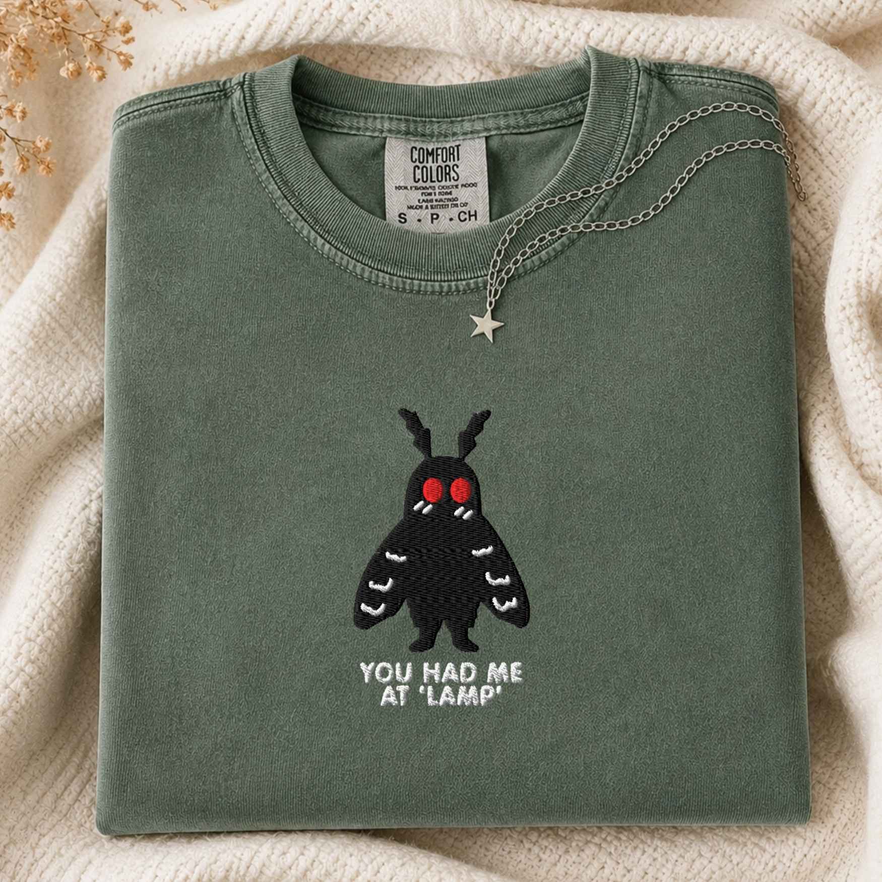 A folded sage green t-shirt with a black moth graphic featuring red eyes and the text 'YOU HAD ME AT "LAMP"' underneath, styled on a cream knit blanket with a silver star necklace.