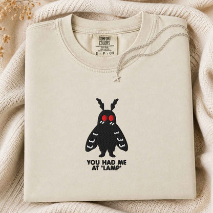 A folded ivory t-shirt with a black moth graphic featuring red eyes and the text 'YOU HAD ME AT "LAMP"' underneath, styled on a cream knit blanket with a silver star necklace.