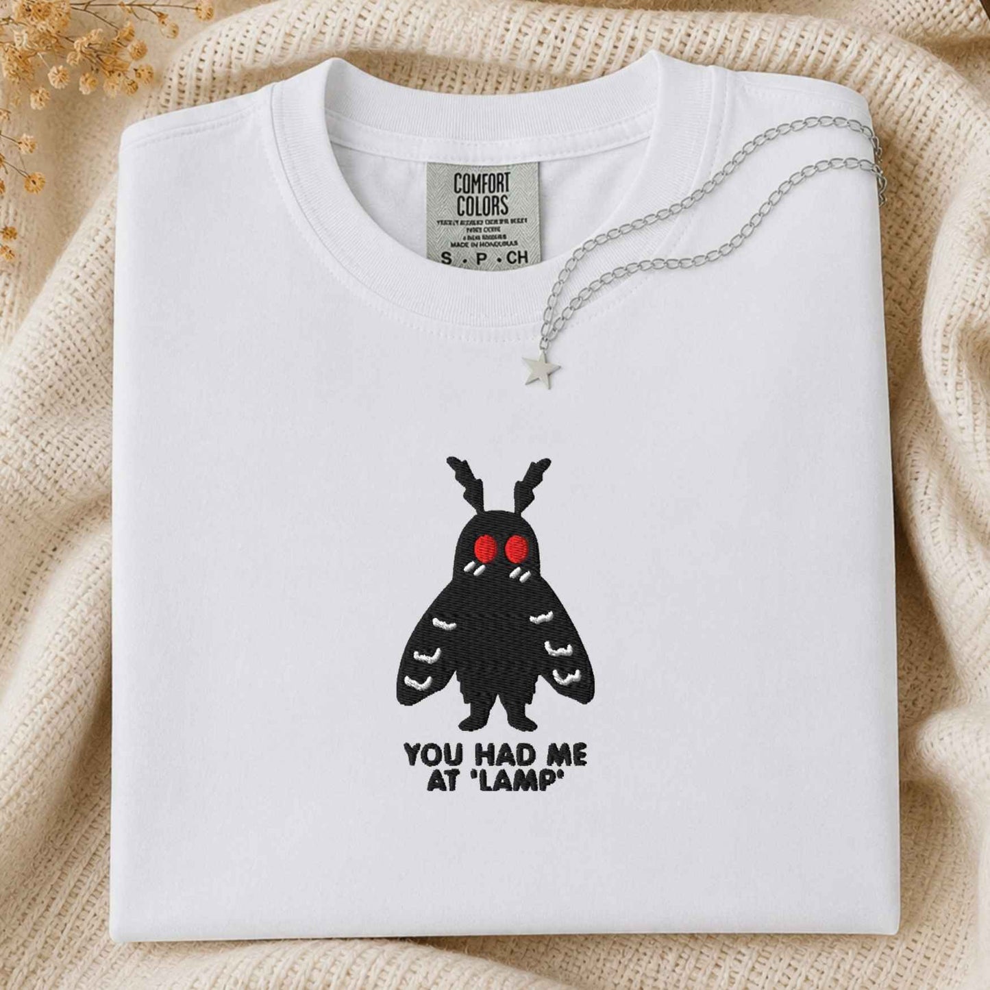 A folded white t-shirt with a black moth graphic featuring red eyes and the text 'YOU HAD ME AT "LAMP"' underneath, styled on a cream knit blanket with a silver star necklace.