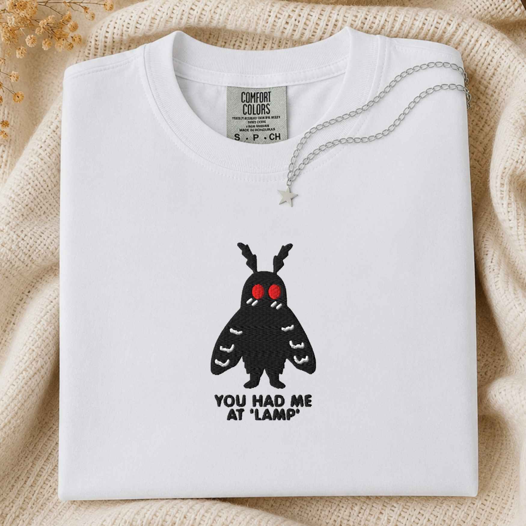 A folded white t-shirt with a black moth graphic featuring red eyes and the text 'YOU HAD ME AT "LAMP"' underneath, styled on a cream knit blanket with a silver star necklace.