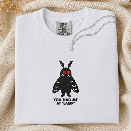 A folded white t-shirt with a black moth graphic featuring red eyes and the text 'YOU HAD ME AT "LAMP"' underneath, styled on a cream knit blanket with a silver star necklace.