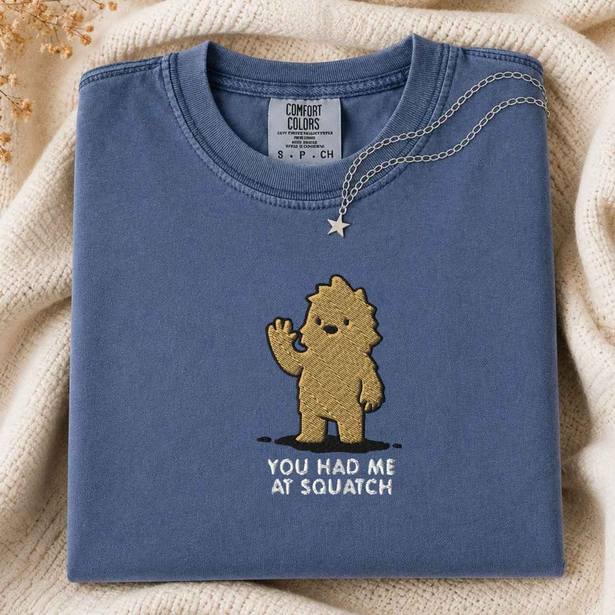 A folded slate blue t-shirt with a gold embroidered cartoon character and the text 'YOU HAD ME AT SQUATCH' on the front, styled on a cream knit blanket with a gold necklace draped over the collar. The neck label shows the brand 'Comfort Colors'.