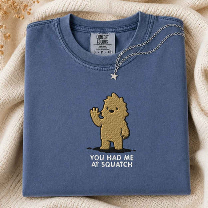 A folded slate blue t-shirt with a gold embroidered cartoon character and the text 'YOU HAD ME AT SQUATCH' on the front, styled on a cream knit blanket with a gold necklace draped over the collar. The neck label shows the brand 'Comfort Colors'.