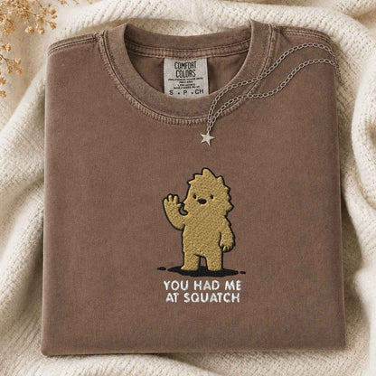 A folded brown tshirt with a gold embroidered cartoon bigfoot and the text 'YOU HAD ME AT SQUATCH' on the front, styled on a cream knit blanket with a silver necklace draped over the collar. The neck label reads 'COMFORT COLORS'.
