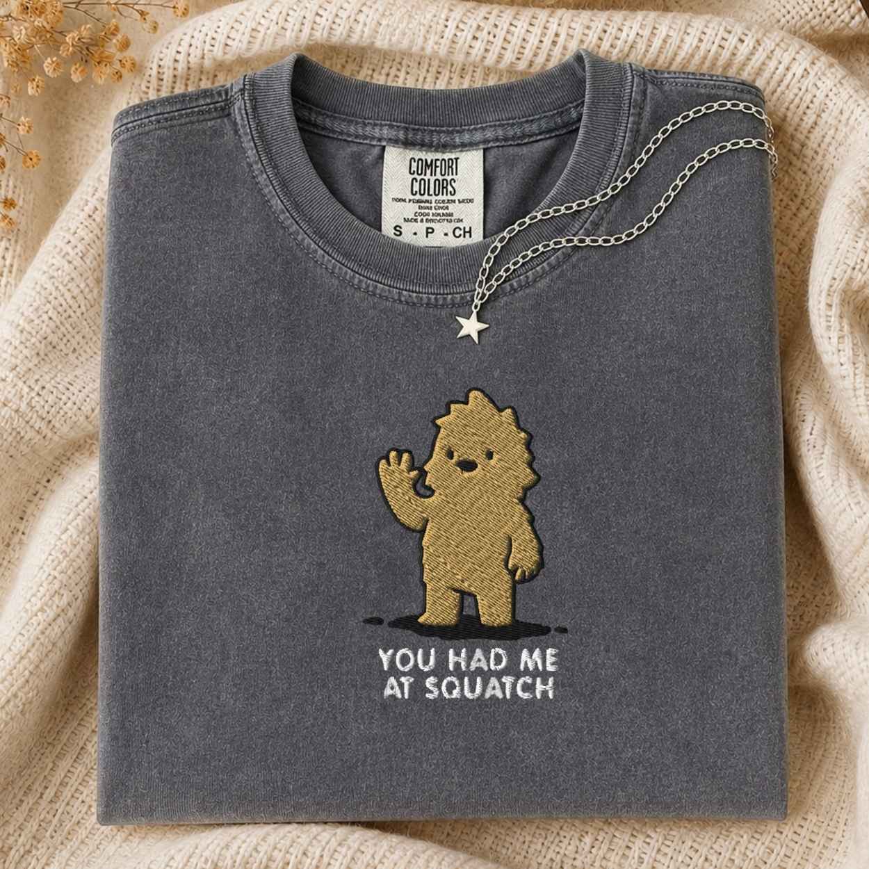 A folded dark gray t-shirt with a brown embroidered cartoon bigfoot and the text 'YOU HAD ME AT SQUATCH' on the front, styled on a beige knit blanket with a silver necklace draped over the collar.