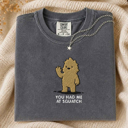 A folded dark gray t-shirt with a brown embroidered cartoon bigfoot and the text 'YOU HAD ME AT SQUATCH' on the front, styled on a beige knit blanket with a silver necklace draped over the collar.