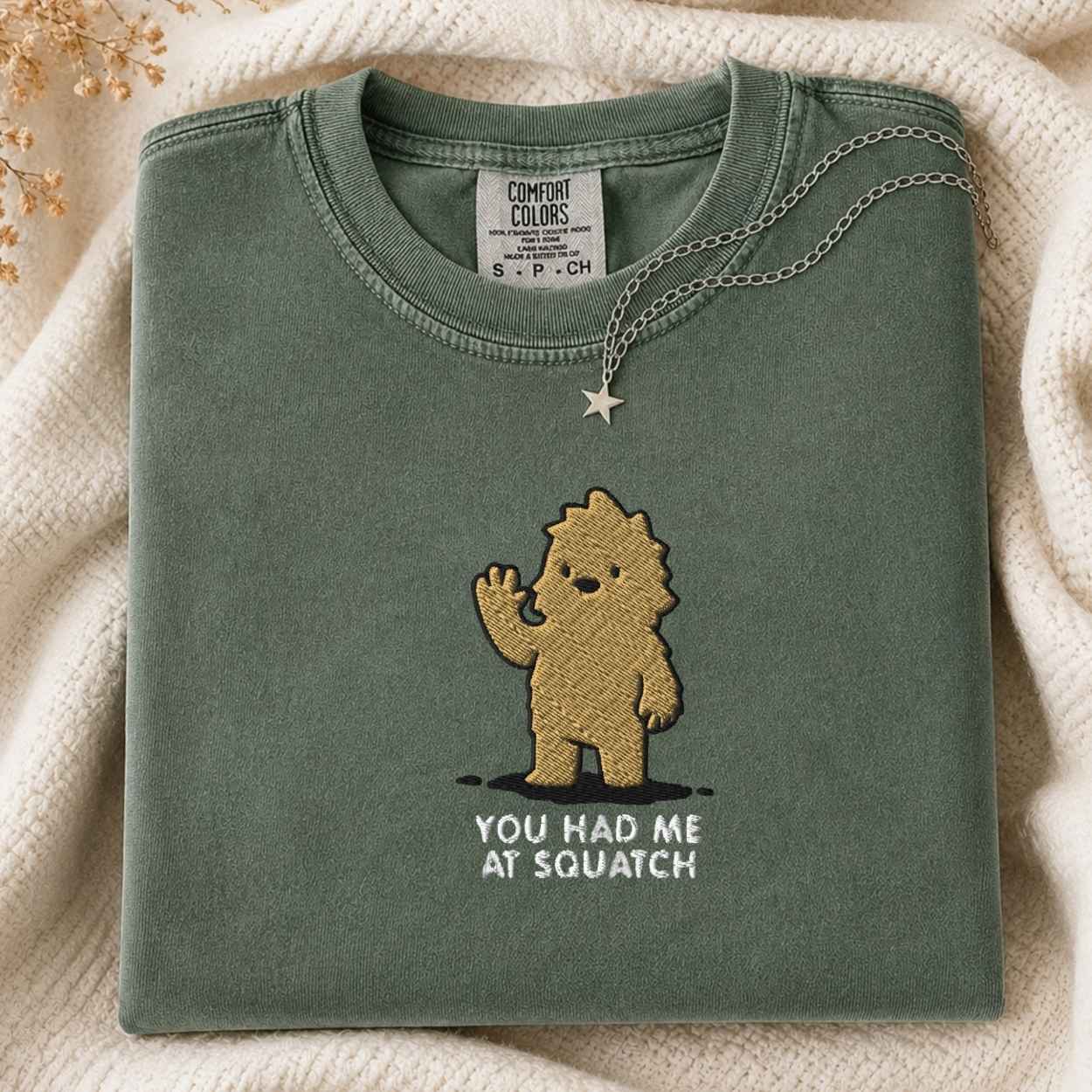 A folded sage green t-shirt with a gold embroidered cartoon character and the text 'YOU HAD ME AT SQUATCH' on the front, styled on a cream knit blanket with a silver necklace.