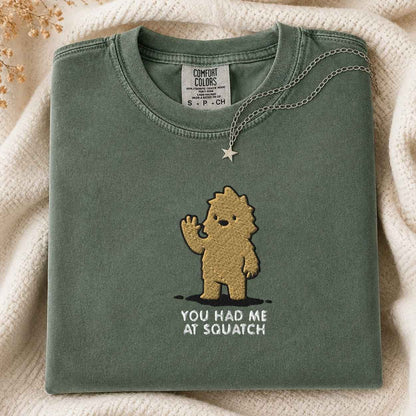 A folded sage green t-shirt with a gold embroidered cartoon character and the text 'YOU HAD ME AT SQUATCH' on the front, styled on a cream knit blanket with a silver necklace.
