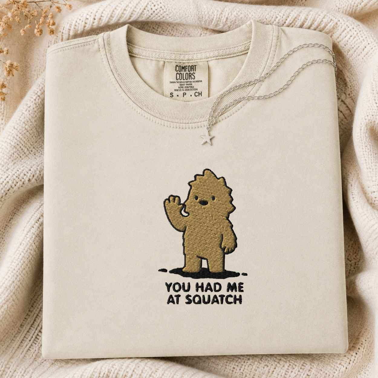 A folded cream-colored tshirt with a graphic of a brown cartoon character waving and the text 'YOU HAD ME AT SQUATCH' underneath, styled on a cozy cream knit blanket.