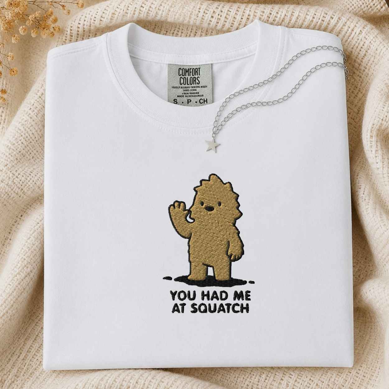 A folded white Comfort Colors t-shirt with a brown embroidered cartoon character and the text 'YOU HAD ME AT SQUATCH' on the front, styled on a beige knit blanket with a silver necklace.