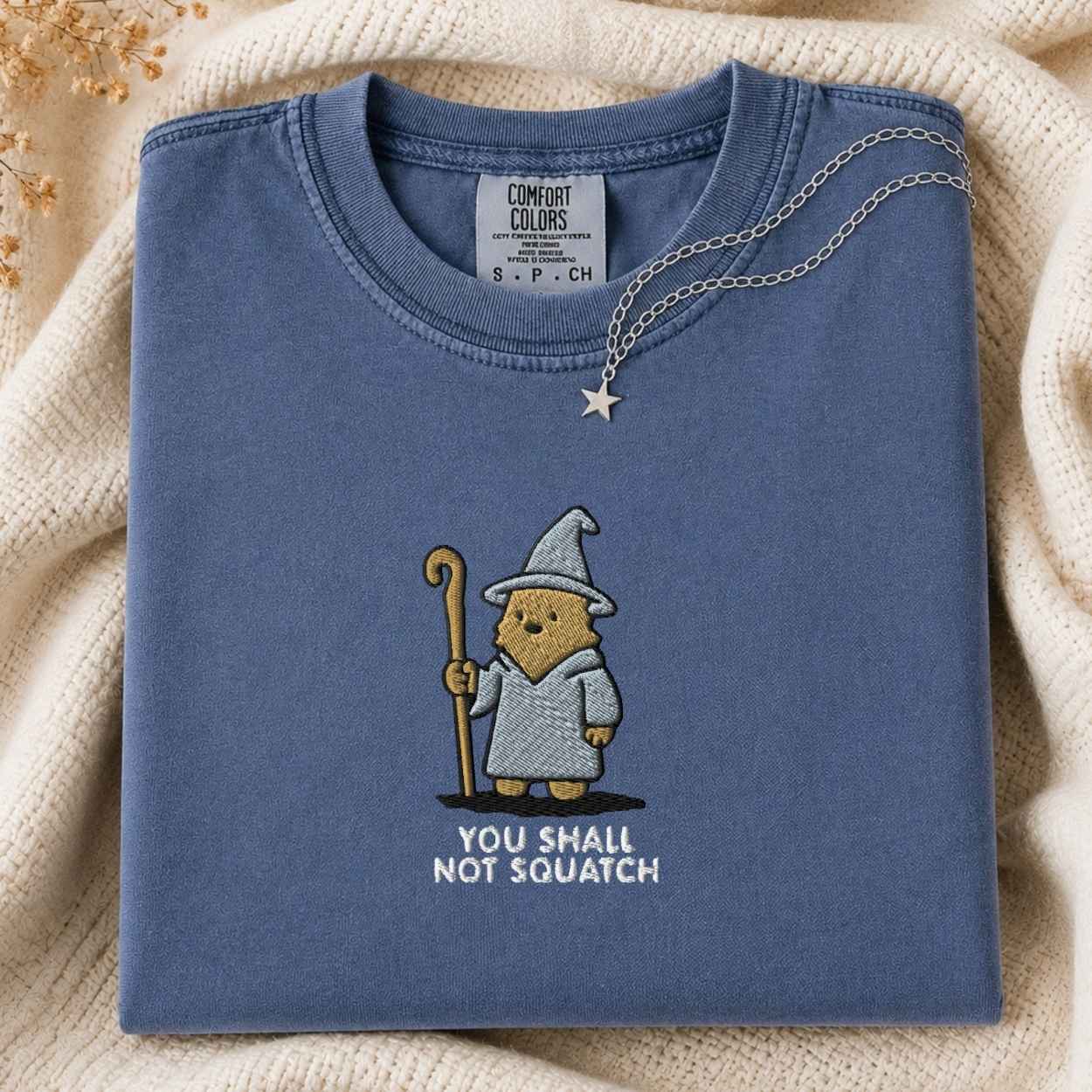 A folded slate blue t-shirt with a squatch character in a wizard hat holding a staff, with the text 'YOU SHALL NOT SQUATCH' underneath. A silver necklace with a star pendant is draped over the collar.