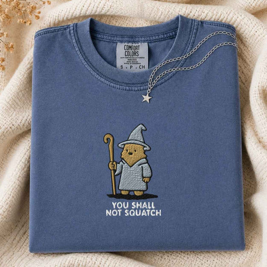 A folded slate blue t-shirt with a squatch character in a wizard hat holding a staff, with the text 'YOU SHALL NOT SQUATCH' underneath. A silver necklace with a star pendant is draped over the collar.