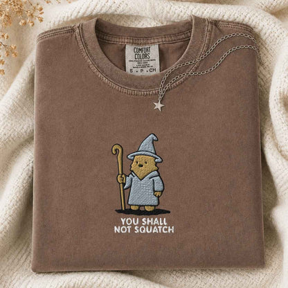 A folded brown sweatshirt with a cartoon bigfoot wearing a wizard hat and robe holding a staff, with the text 'YOU SHALL NOT SQUATCH' below. A silver star pendant necklace is draped over the collar.