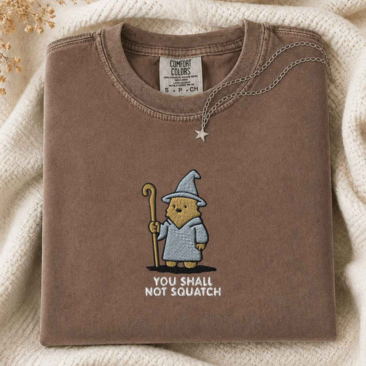 A folded brown sweatshirt with a cartoon bigfoot wearing a wizard hat and robe holding a staff, with the text 'YOU SHALL NOT SQUATCH' below. A silver star pendant necklace is draped over the collar.