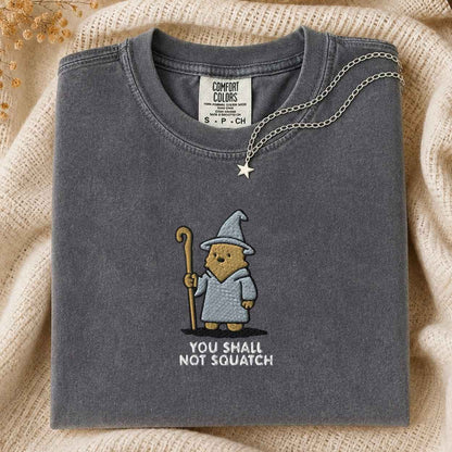 A folded dark gray t-shirt with a bigfoot wearing a wizard hat and robe holding a staff, with the text 'YOU SHALL NOT SQUATCH' underneath. A silver star necklace is draped over the collar.