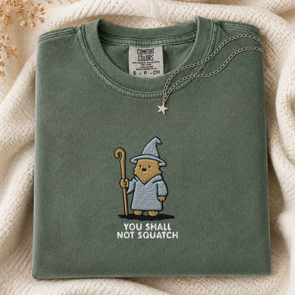 A folded sage green Comfort Colors t-shirt with a cute embroidered wizard squatch holding a staff and the text 'YOU SHALL NOT SQUATCH'. The shirt is styled on a cream knit blanket with a silver star necklace draped over the collar.