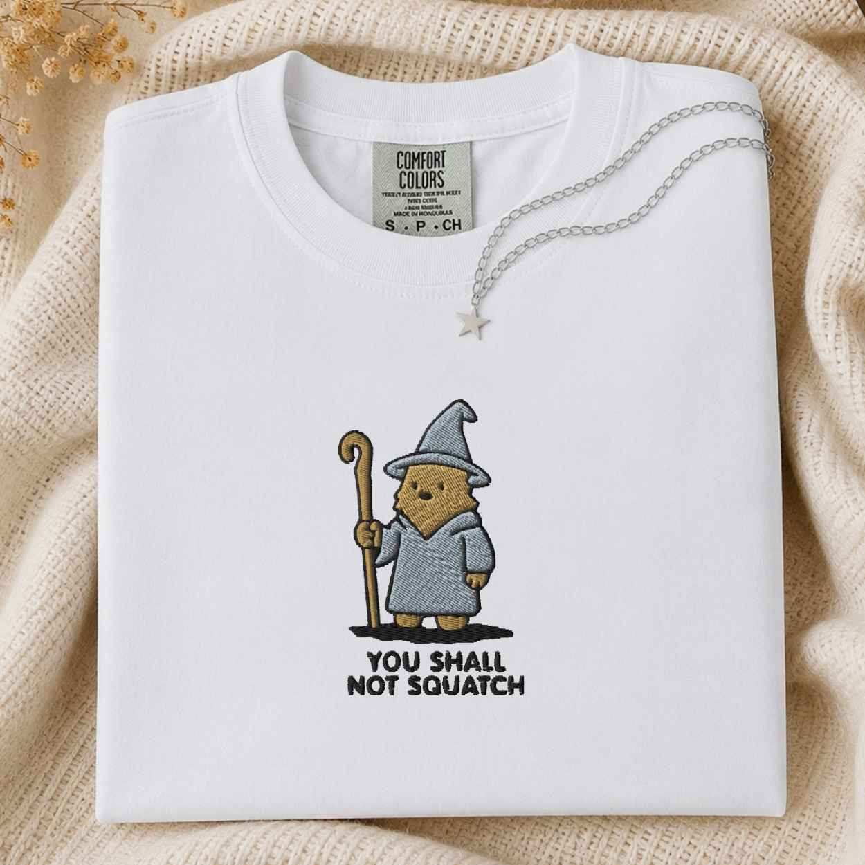 A folded white t-shirt with a cartoon wizard bigfoot graphic and the text 'YOU SHALL NOT SQUATCH' on the front, styled on a beige knit blanket with a silver necklace.