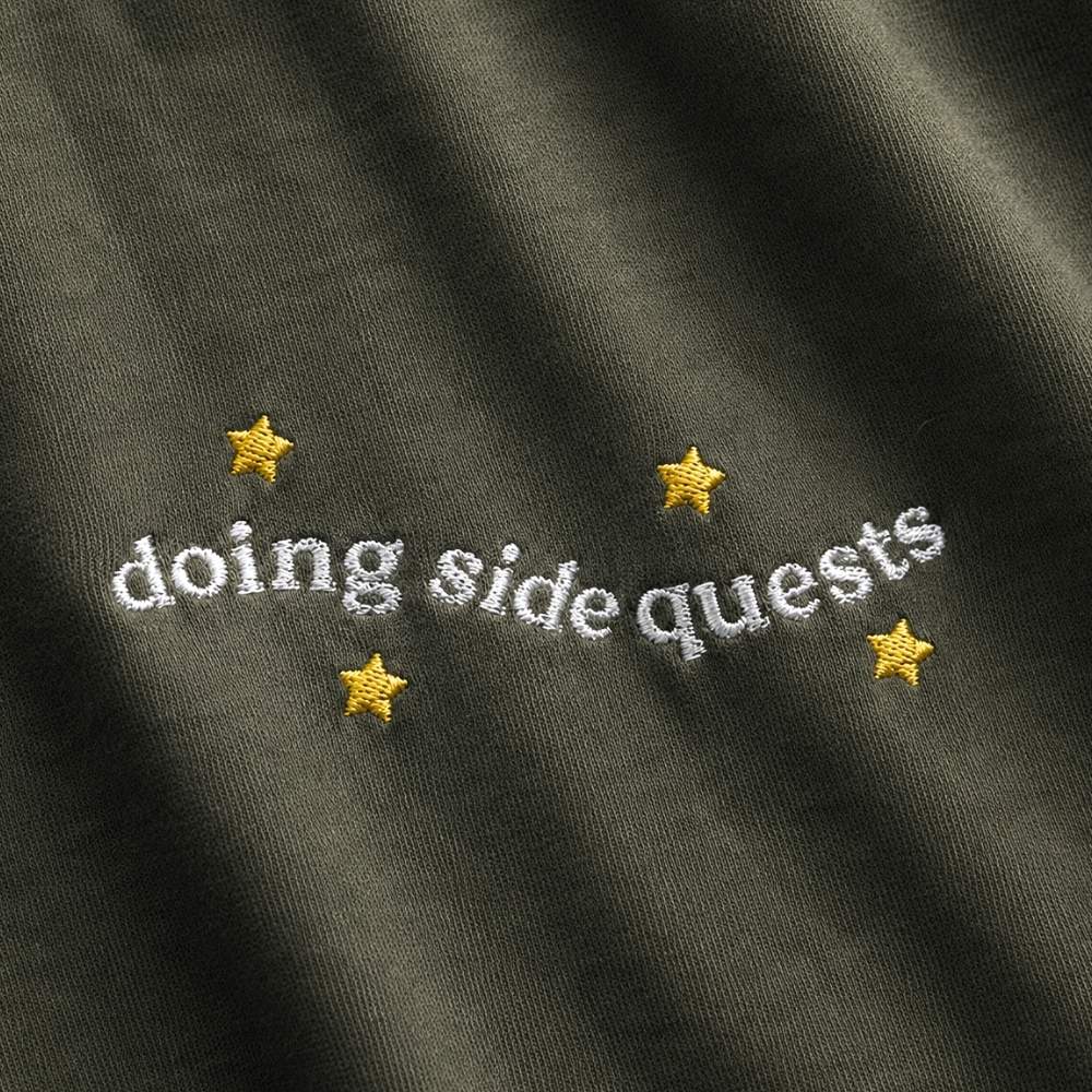 Close-up of a dark olive green fabric surface with embroidered white text reading 'doing side quests' and four small gold stars.