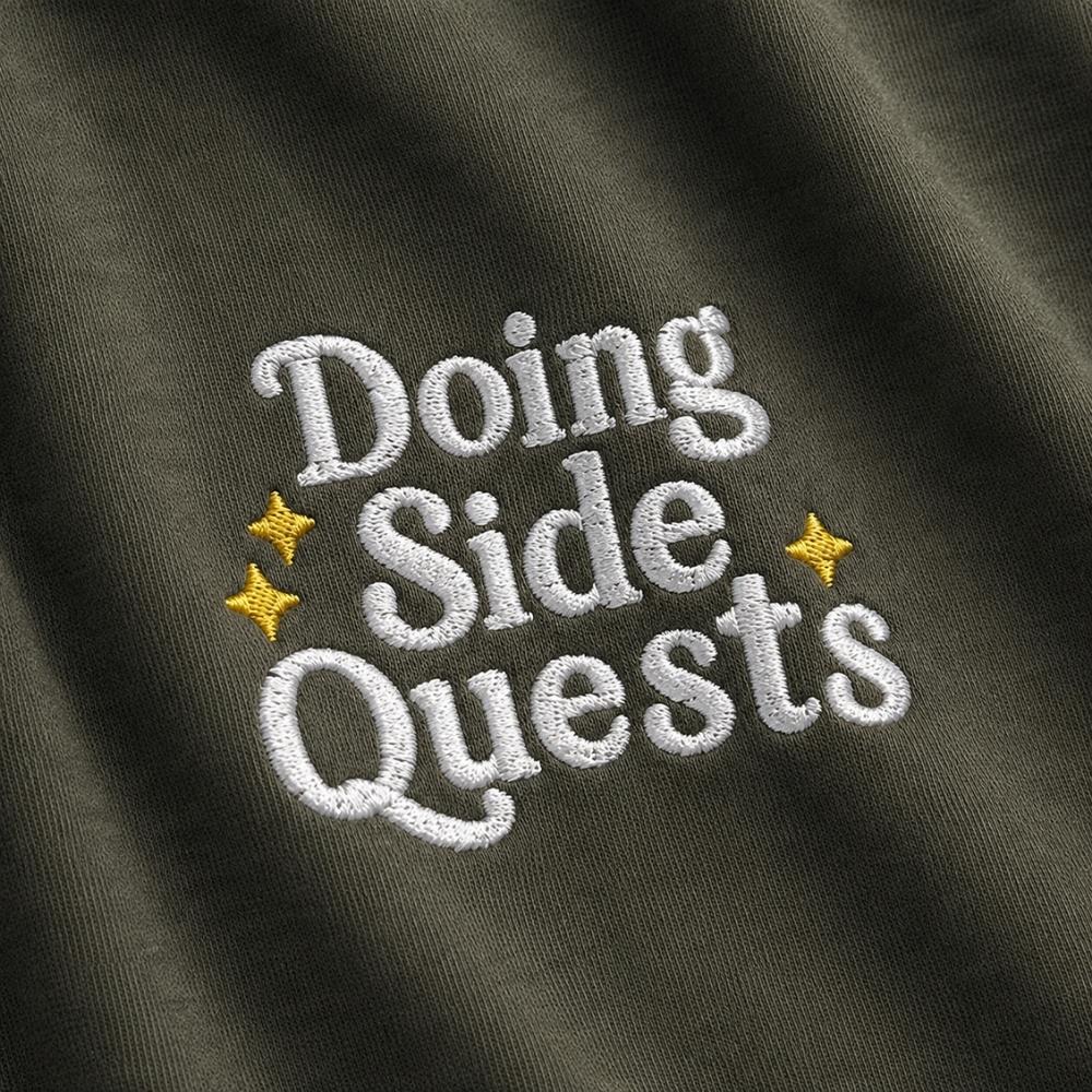 Close-up of embroidered text reading 'Doing Side Quests' in white on an olive green fabric, with two small yellow star accents.