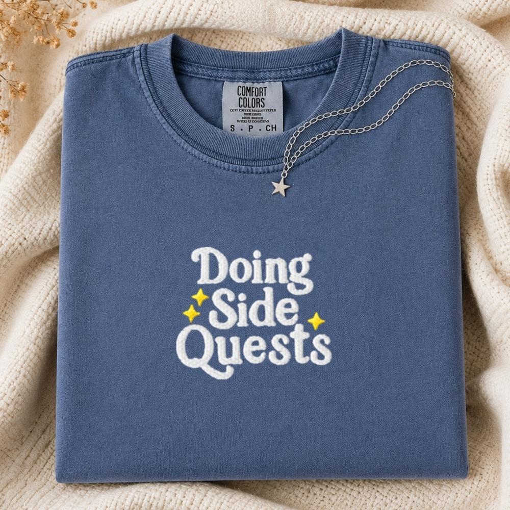 A folded slate blue t-shirt with 'Doing Side Quests' printed in white and yellow on the front, styled on a cream knit blanket with a silver necklace.