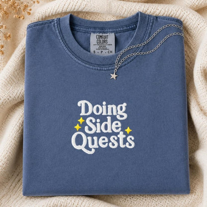 A folded slate blue t-shirt with 'Doing Side Quests' printed in white and yellow on the front, styled on a cream knit blanket with a silver necklace.