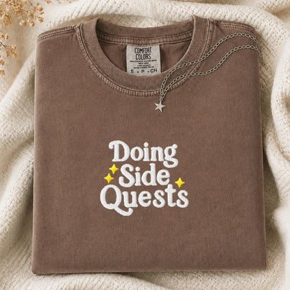 A folded brown Comfort Colors t-shirt with 'Doing Side Quests' printed in white and yellow, styled on a cream knit blanket with a silver necklace.