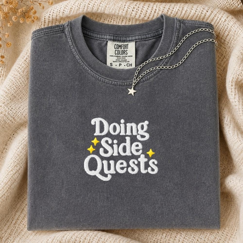 A folded dark gray Comfort Colors t-shirt with 'Doing Side Quests' printed in white and yellow, styled on a beige knit blanket with a silver star necklace.