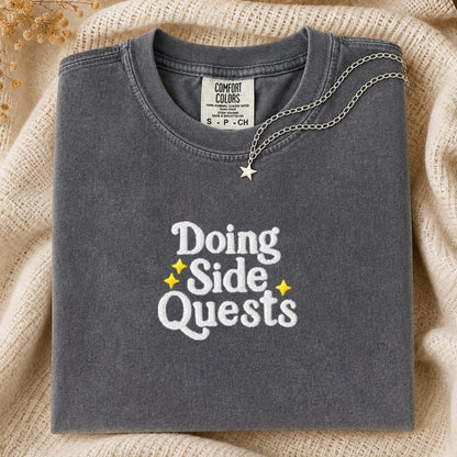A folded dark gray Comfort Colors t-shirt with 'Doing Side Quests' printed in white and yellow, styled on a beige knit blanket with a silver star necklace.