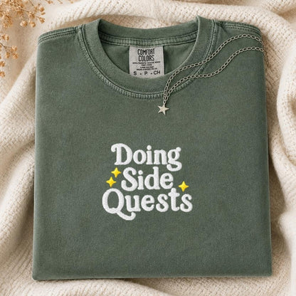 A folded sage green Comfort Colors t-shirt with 'Doing Side Quests' printed in white and yellow on the front, styled on a cream knit blanket with a silver necklace.
