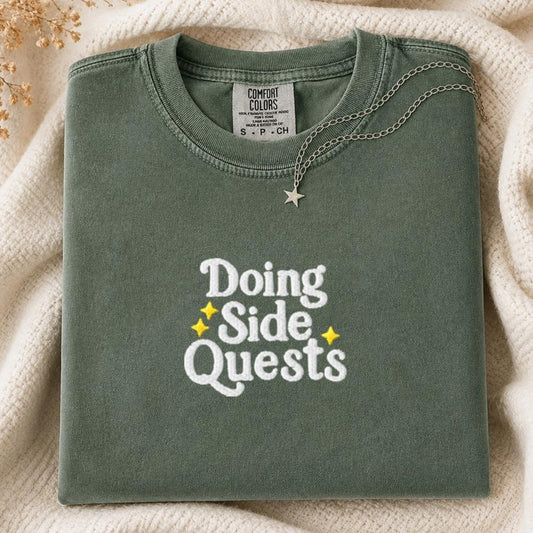 A folded sage green Comfort Colors t-shirt with 'Doing Side Quests' printed in white and yellow on the front, styled on a cream knit blanket with a silver necklace.