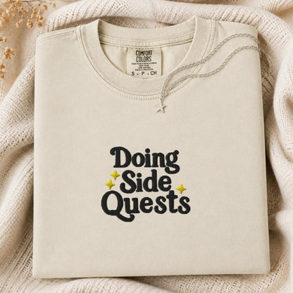 A folded cream-colored crewneck tshirt with the text 'Doing Side Quests' printed in black with small yellow star accents, displayed on a cozy cream knit blanket.