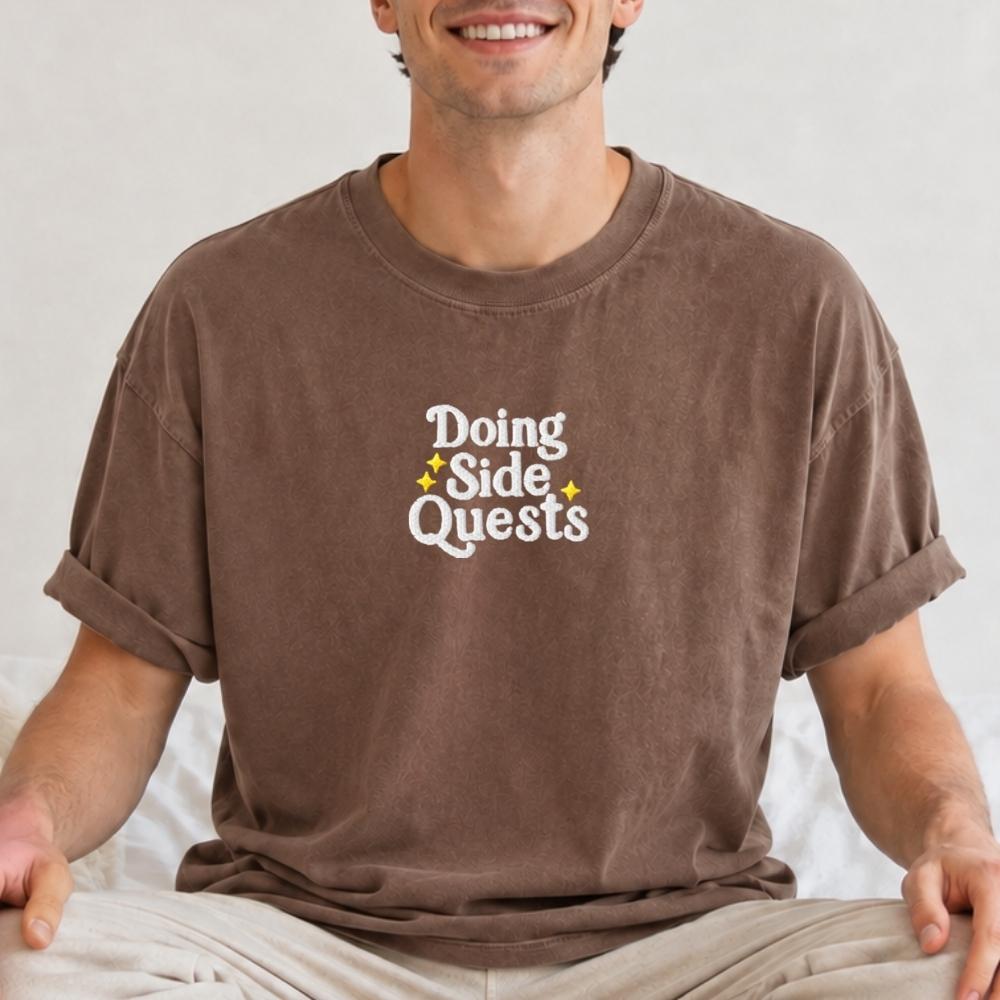 A man wearing a brown t-shirt with rolled-up sleeves and light beige pants, sitting cross-legged. The t-shirt features a white graphic text reading 'Doing Side Quests' with small yellow star accents.