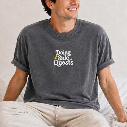 A man wearing a dark gray washed t-shirt with the text 'Doing Side Quests' in white and small yellow star accents, paired with light beige pants, sitting on a bed.