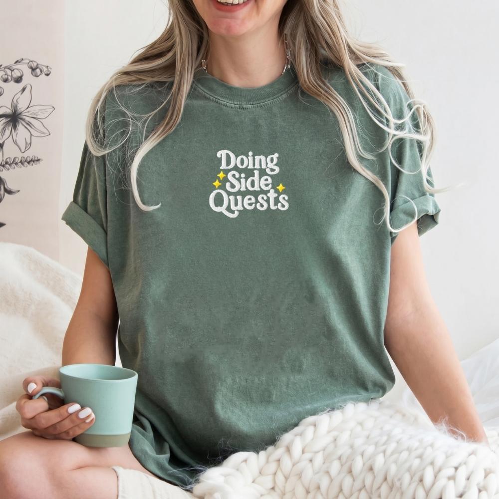 A woman wearing a sage green t-shirt with the text 'Doing Side Quests' in white with small yellow star accents, holding a mint green mug while sitting on a bed with a chunky knit blanket.