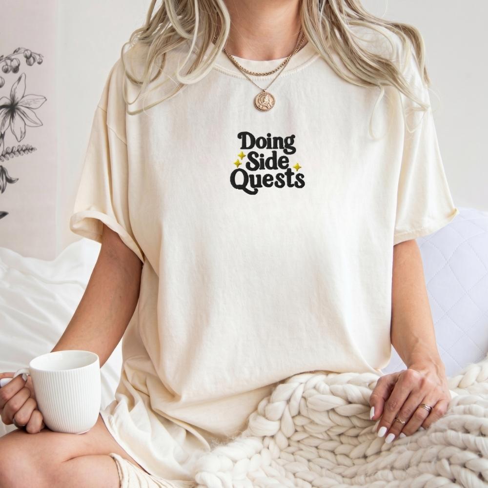 A woman wearing an oversized cream-colored t-shirt with the text 'Doing Side Quests' printed in black with small yellow star accents. She is holding a white mug and sitting on a chunky knit blanket.