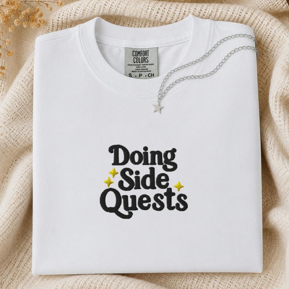 A folded white Comfort Colors t-shirt with 'Doing Side Quests' printed in black text with small yellow star accents, styled on a beige knit blanket with a silver necklace draped over it.