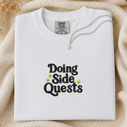 A folded white Comfort Colors t-shirt with 'Doing Side Quests' printed in black text with small yellow star accents, styled on a beige knit blanket with a silver necklace draped over it.