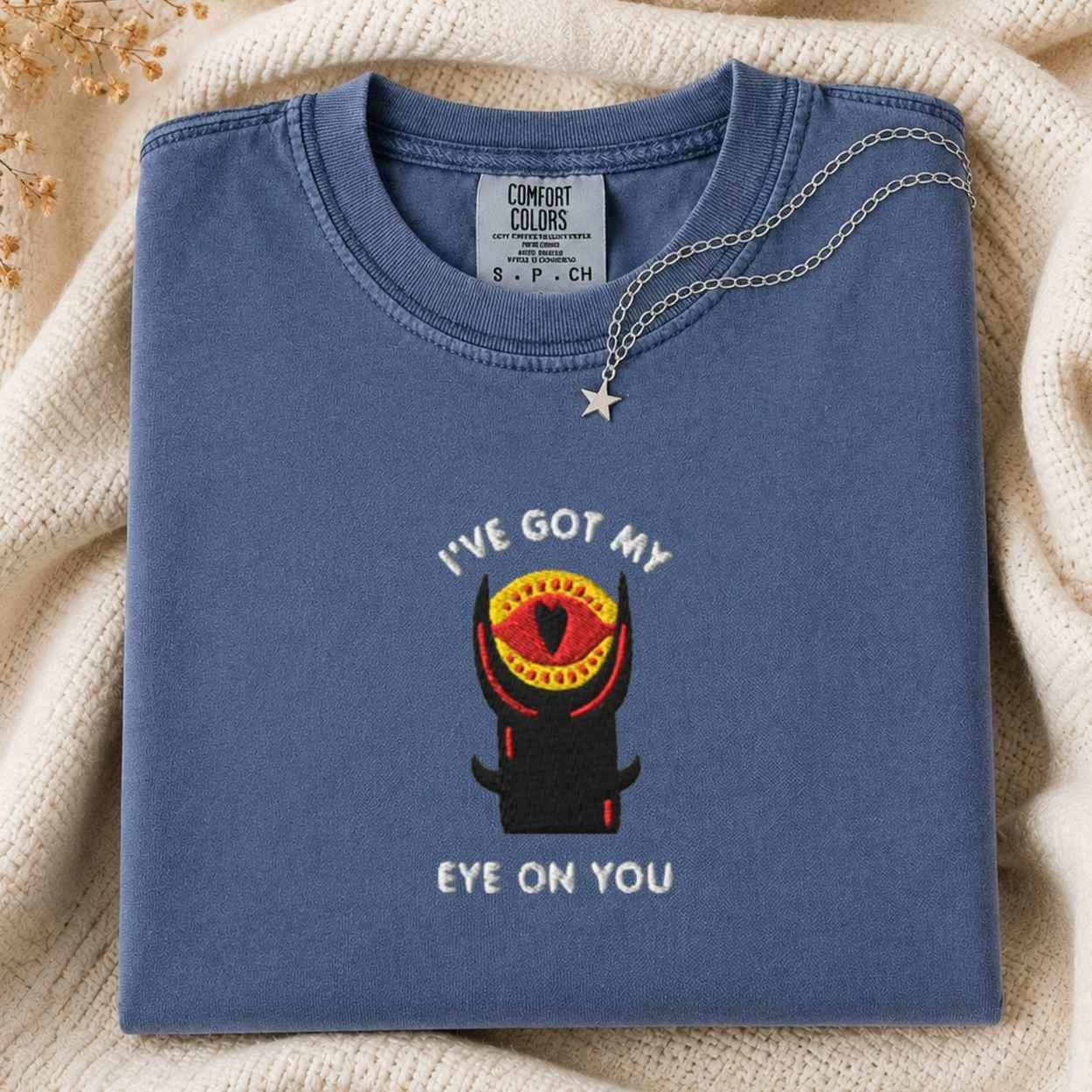 A folded slate blue t-shirt with a graphic of a tower with a single red eye and the text 'I'VE GOT MY EYE ON YOU'. A silver necklace with a star pendant is draped over the collar.