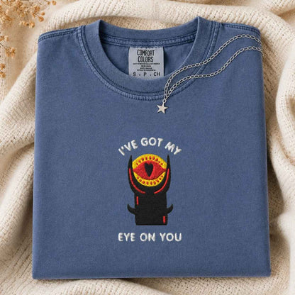 A folded slate blue t-shirt with a graphic of a tower with a single red eye and the text 'I'VE GOT MY EYE ON YOU'. A silver necklace with a star pendant is draped over the collar.