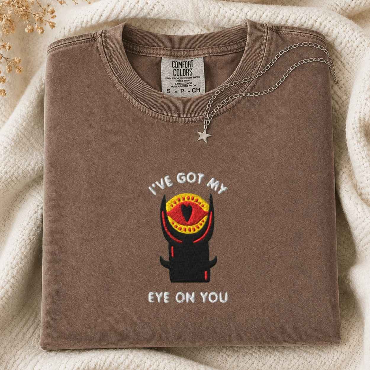 A folded brown Comfort Colors t-shirt with a graphic of a tower with an eye and the text 'I'VE GOT MY EYE ON YOU', styled on a cream knit blanket with a silver necklace.