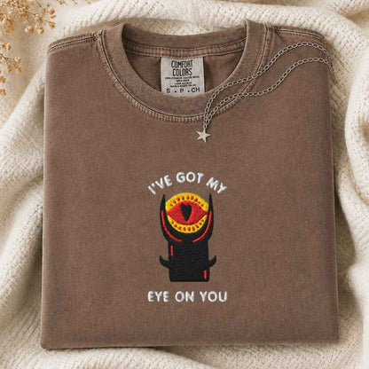 A folded brown Comfort Colors t-shirt with a graphic of a tower with an eye and the text 'I'VE GOT MY EYE ON YOU', styled on a cream knit blanket with a silver necklace.