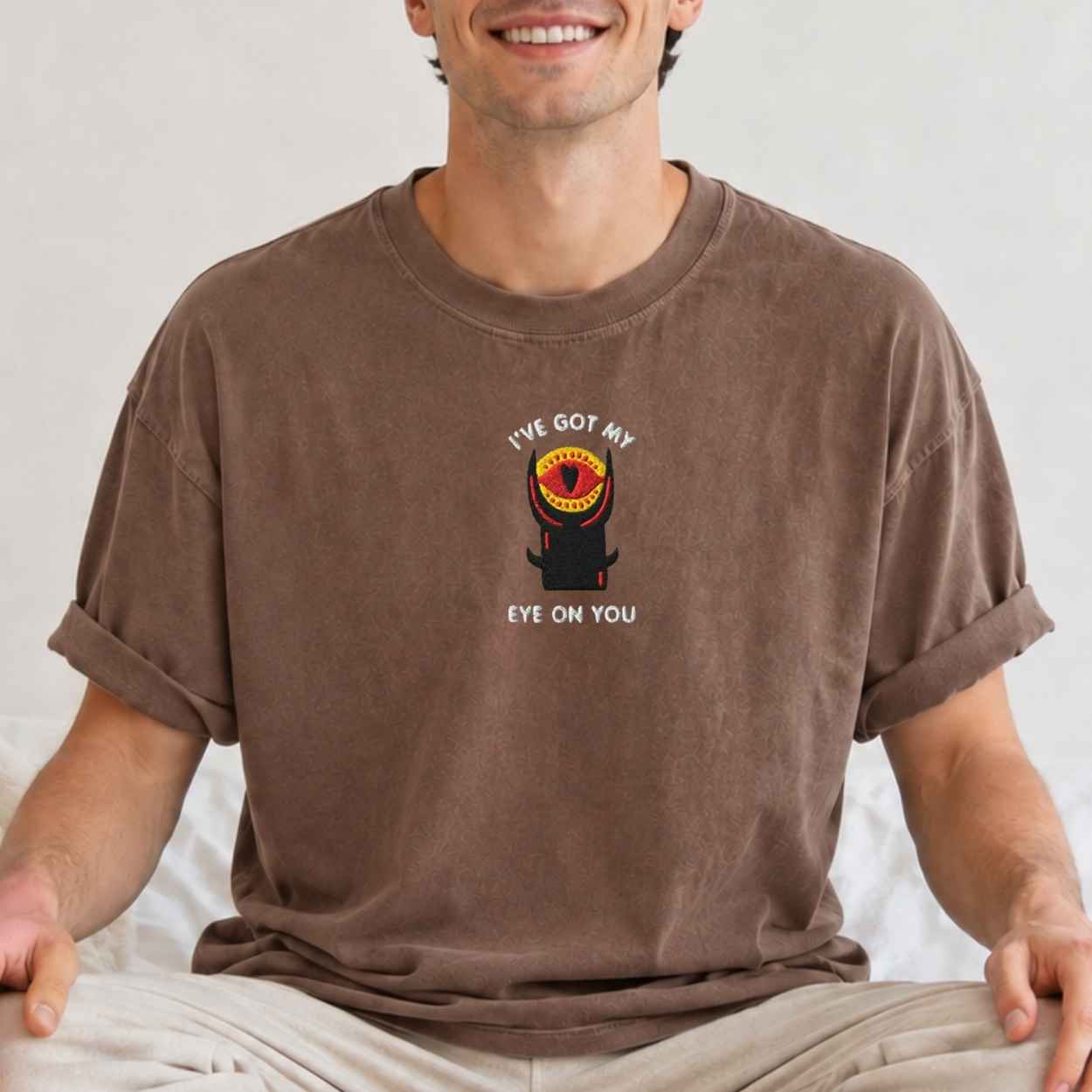 A man wearing a brown crewneck t-shirt with a graphic of a black tower with a red and yellow eye and the text 'I'VE GOT MY EYE ON YOU'.