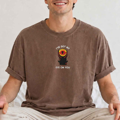 A man wearing a brown crewneck t-shirt with a graphic of a black tower with a red and yellow eye and the text 'I'VE GOT MY EYE ON YOU'.