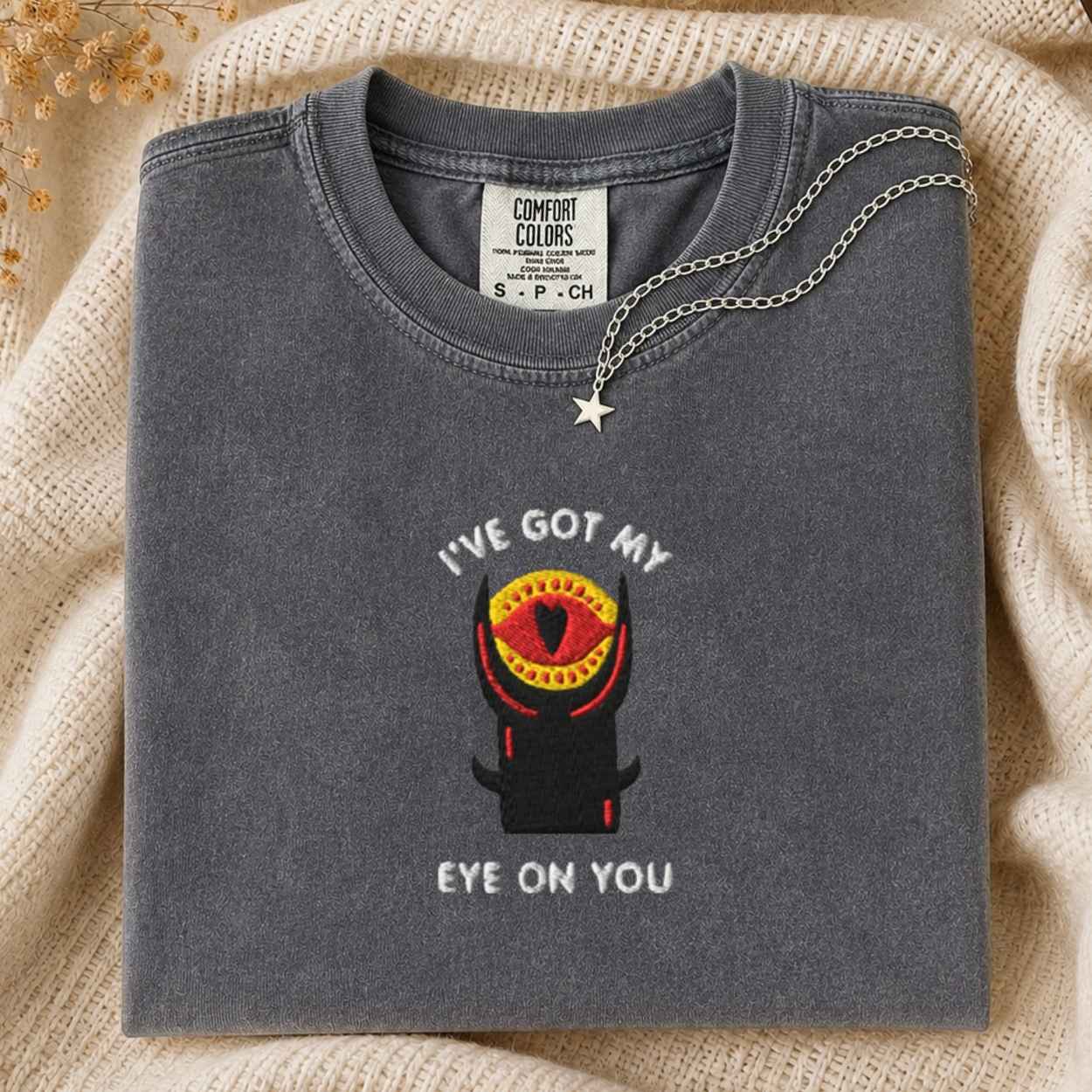 A folded dark gray t-shirt with a graphic of a black tower with a red and yellow eye and the text 'I'VE GOT MY EYE ON YOU'. A silver chain necklace with a star pendant is draped over the collar.
