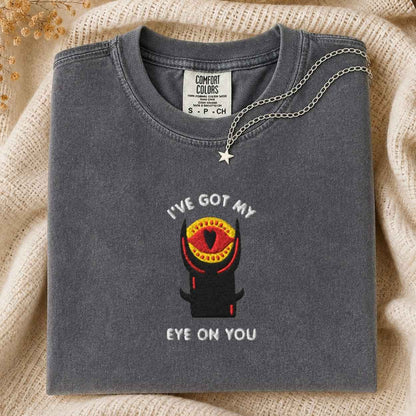 A folded dark gray t-shirt with a graphic of a black tower with a red and yellow eye and the text 'I'VE GOT MY EYE ON YOU'. A silver chain necklace with a star pendant is draped over the collar.
