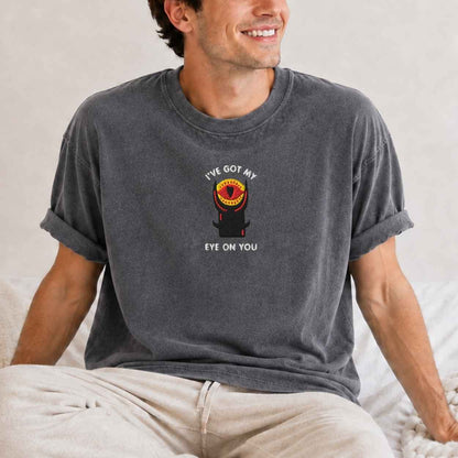 A man wearing a dark gray graphic t-shirt with a tower design and the text 'I'VE GOT MY EYE ON YOU', paired with beige pants, sitting on a bed.