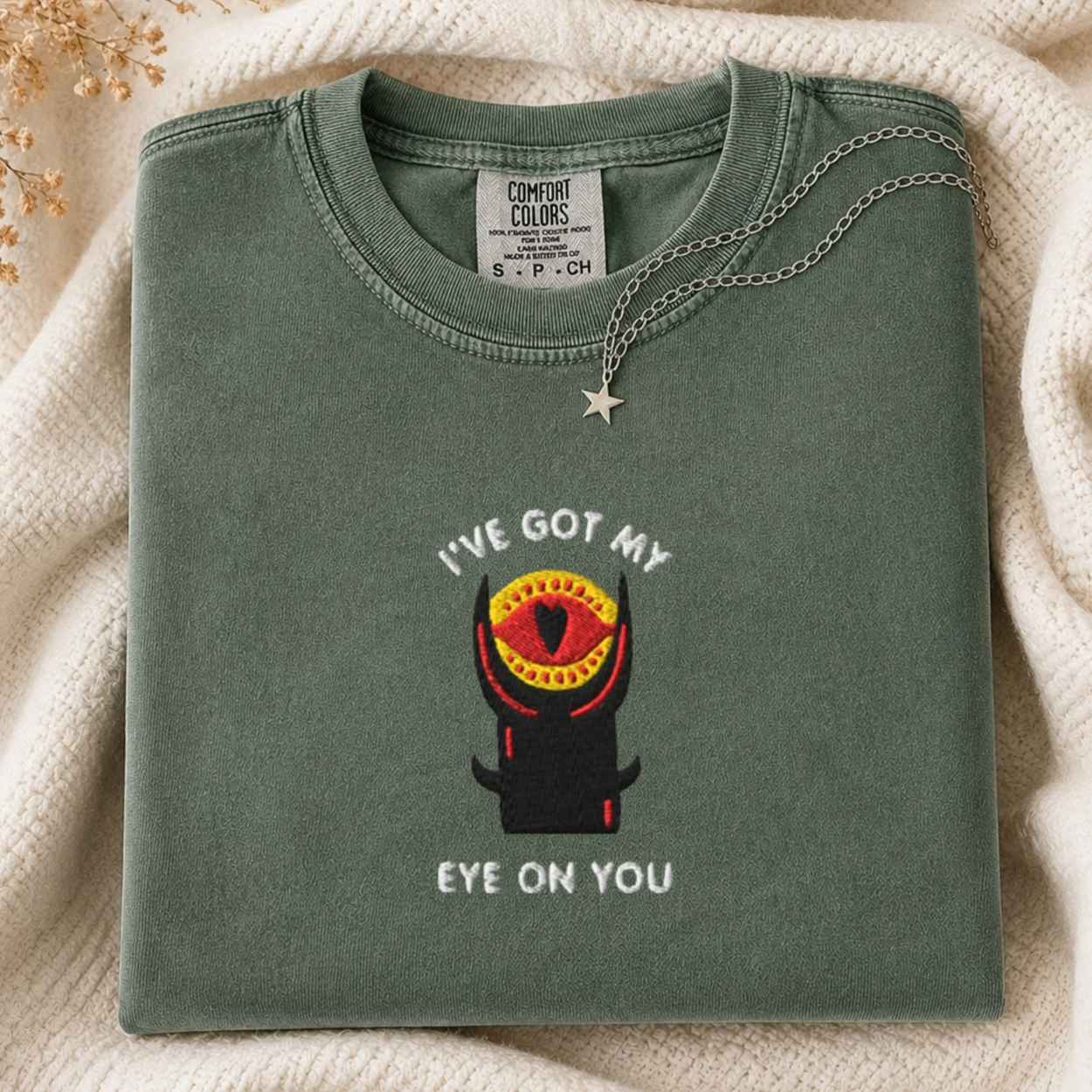 A folded sage green Comfort Colors t-shirt with a graphic of a horned tower with a red heart-shaped eye and the text 'I'VE GOT MY EYE ON YOU'. A silver star necklace is draped over the collar, set against a cozy cream knit blanket background.